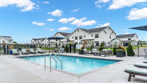 A swimming pool surrounded by lounge chairs and a fence with houses in the background.