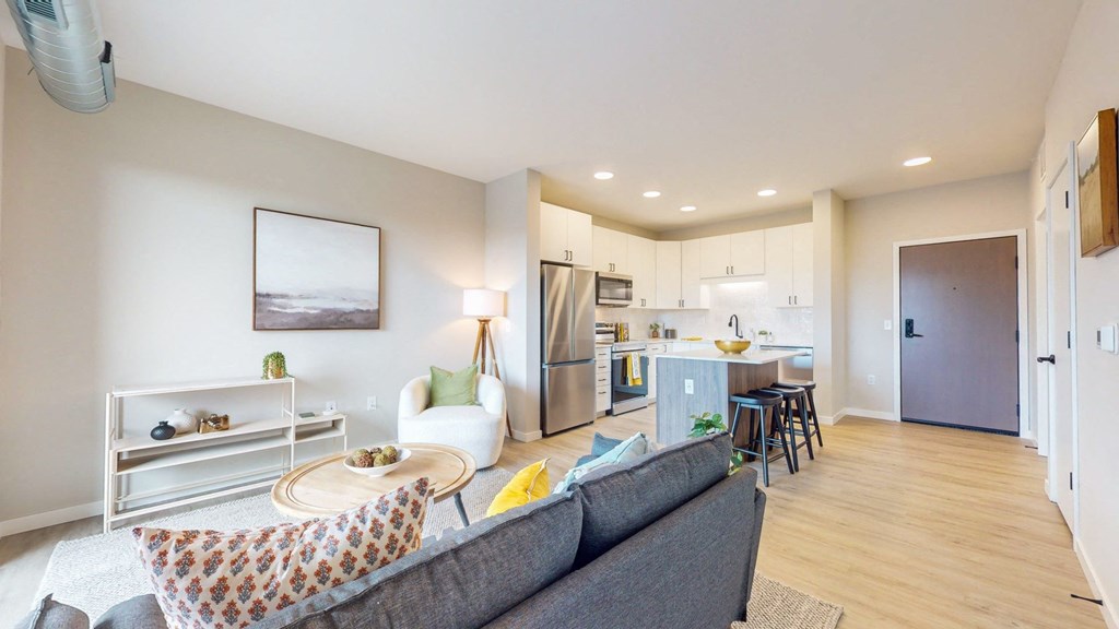 a living room with a couch a table and a chair and a kitchen with a refrigerator and