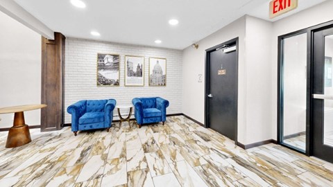 A hallway with a marble floor, two blue chairs, a wooden table, and pictures on the wall.