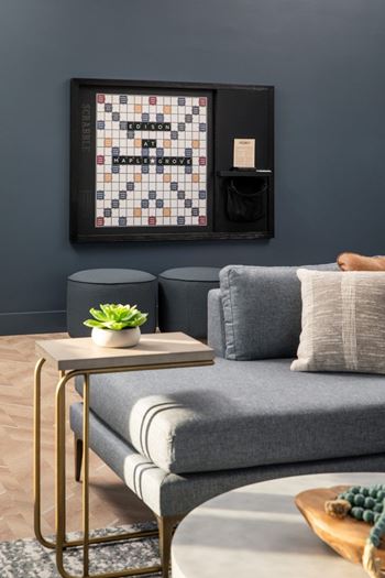 A living room with a grey couch, a small table with a plant and a book, and a framed board game on the wall.at The Edison at Maple Grove, Minnesota