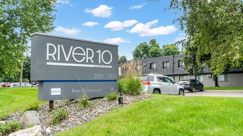 A sign for River10 is in front of a grassy area and some trees.