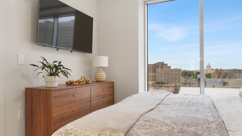 A bedroom with a large bed, a wooden dresser, and a view of the city through a large window.