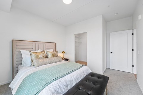 A bedroom with a bed, a black ottoman, and a white door.