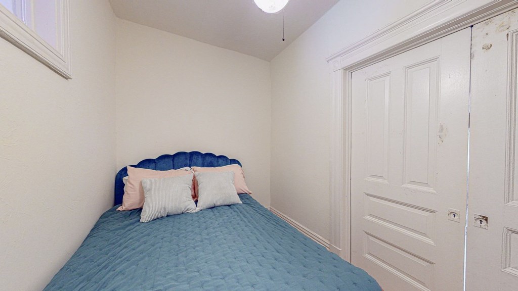 A bed with a blue cover and two pillows sits in a room with white walls.