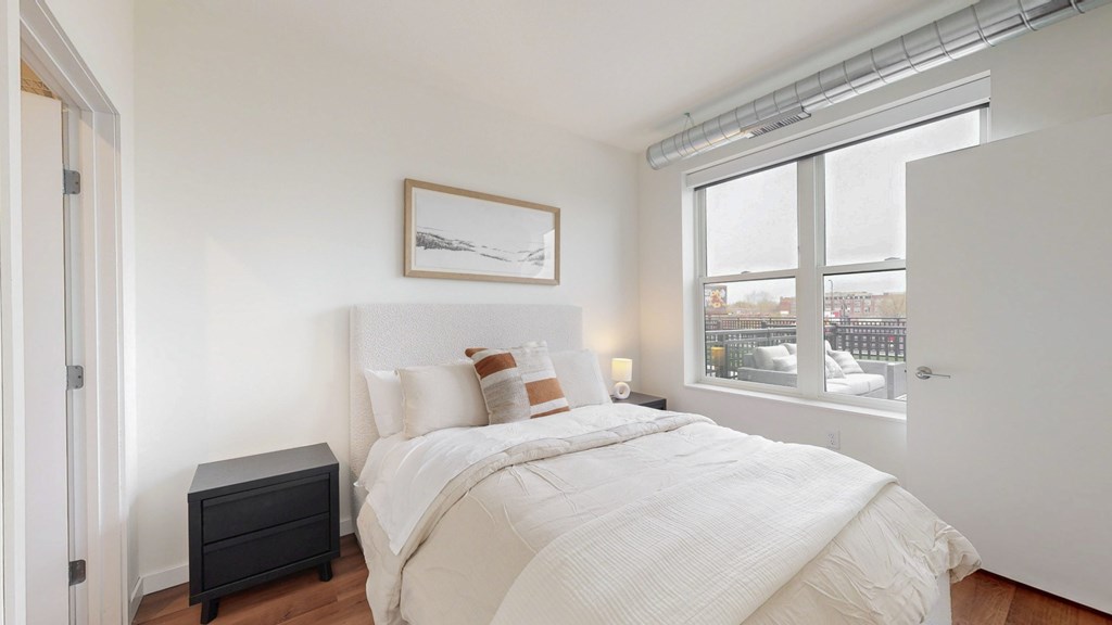 A bedroom with a large bed, a nightstand, and a window overlooking a cityscape.