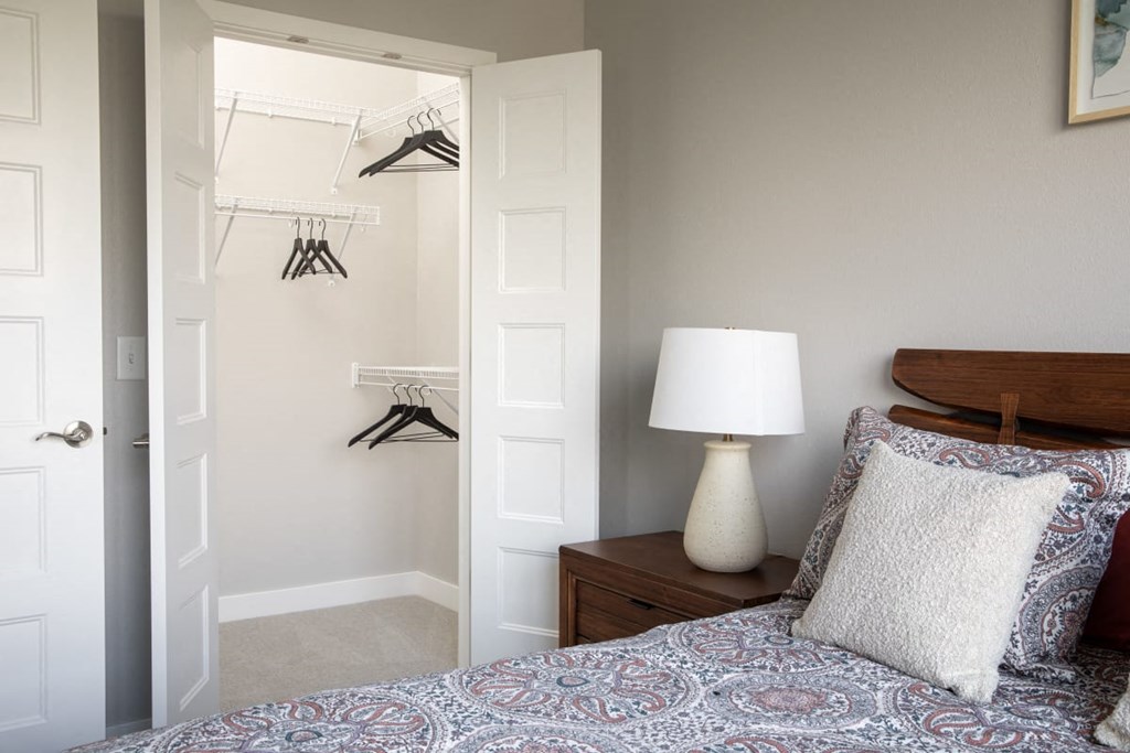 A bedroom with a bed, a nightstand with a lamp, and a closet with clothes hanging.at The Edison at Maple Grove, Maple Grove, MN 55311