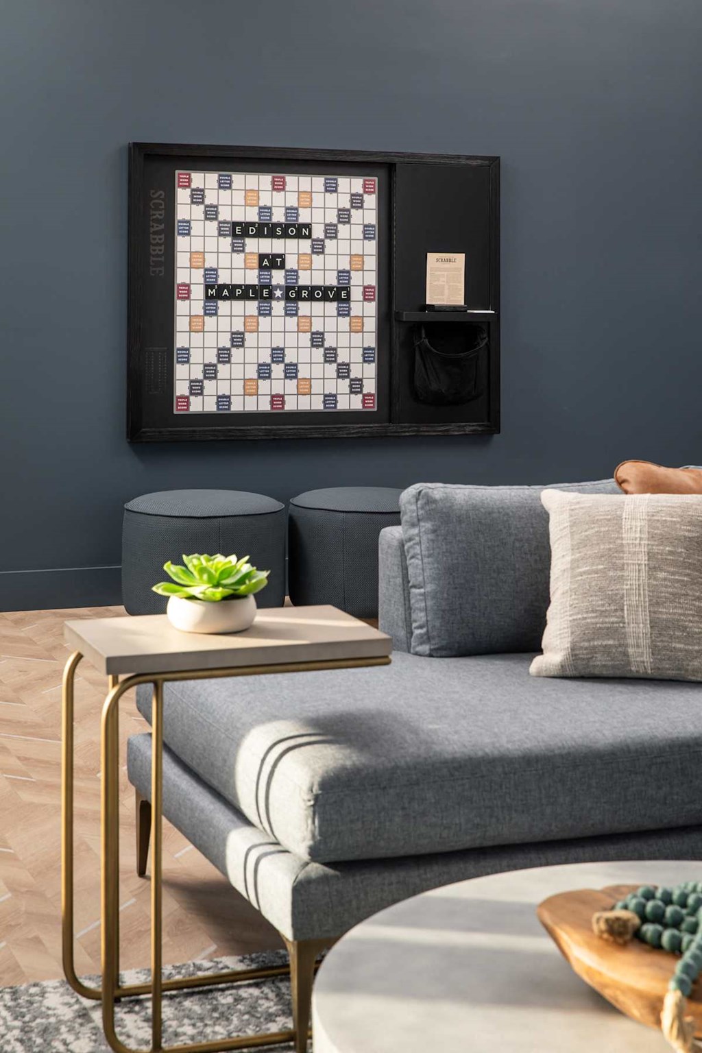 A living room with a grey couch, a small table with a plant and a book, and a framed board game on the wall.at The Edison at Maple Grove, Minnesota, 55311