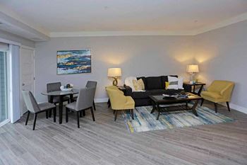 A living room with a grey couch, a yellow chair, a black chair, a coffee table, and a painting on the wall at The Edison at Spirit, Minnesota