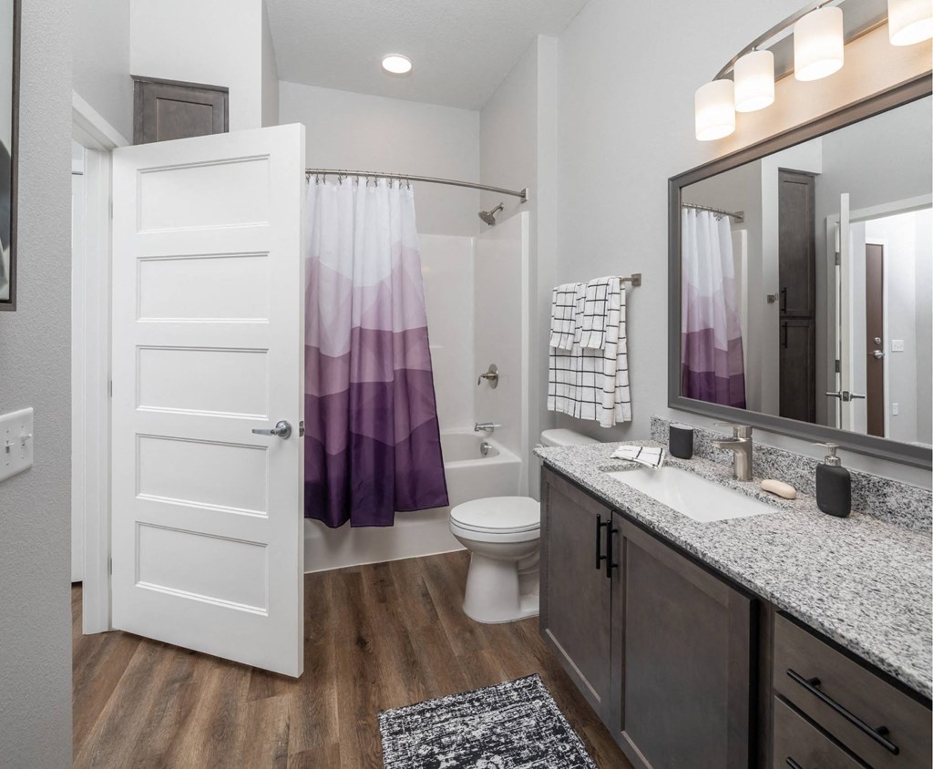 A bathroom with a white door, a purple shower curtain, and a grey granite countertop at The Edison at Woodbury, Woodbury, MN 55129