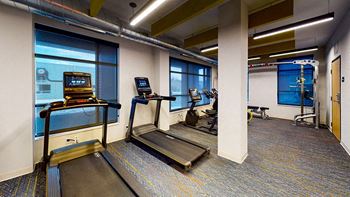 A treadmill and a stationary bike are in a well-lit room with a view of the outside at The Bessemer at Seward Commons Apartments, Minnesota