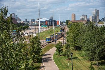 A train is passing by a park with a city skyline in the background at The Bessemer at Seward Commons Apartments, Minneapolis, 55404