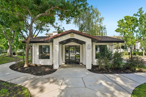 A small building with a brown roof and white walls is surrounded by trees and shrubs.