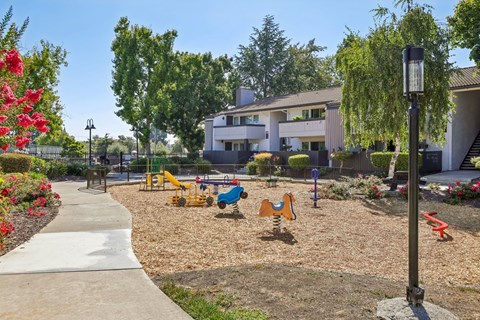 A playground with a slide, swings, and a climbing frame is surrounded by trees and flowers.