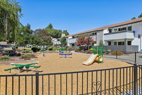 A playground with a slide, swings, and a seesaw in front of a building.