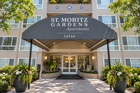 The front entrance of the St. Moritz Gardens Apartments is welcoming with a black awning and two large planters with flowers.