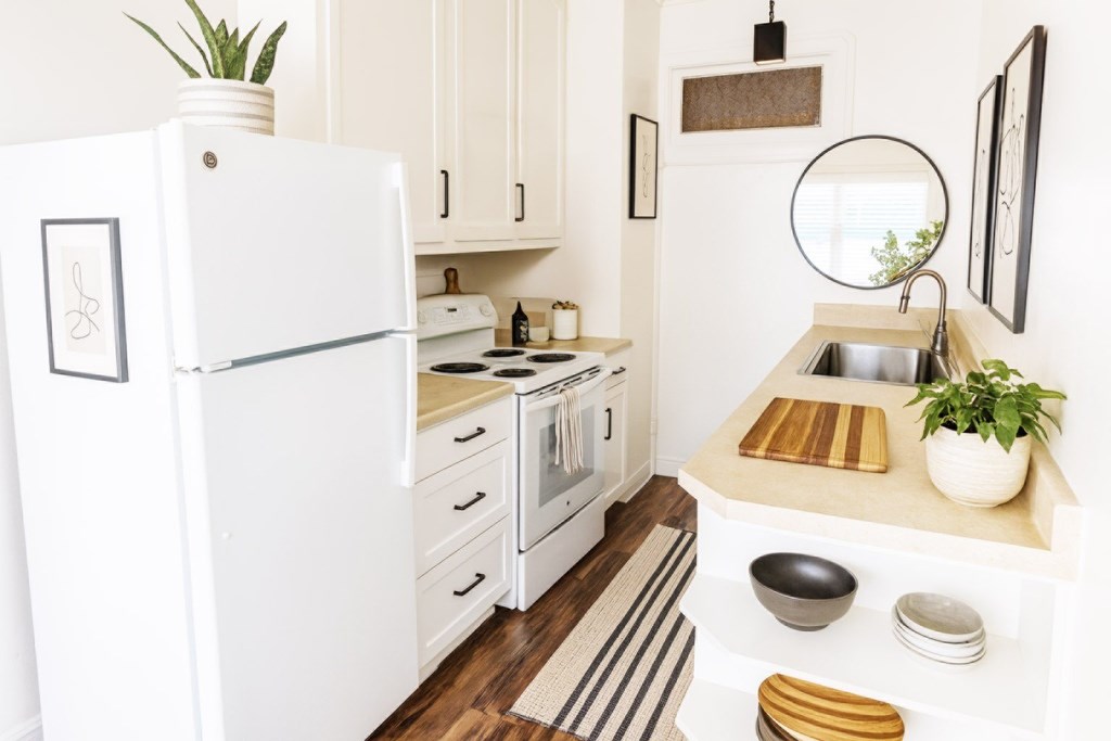 A white refrigerator stands in a kitchen with a sink and a mirror.