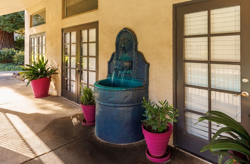 A blue fountain is in the middle of a concrete area with potted plants on each side.