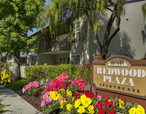 A sign for Redwood Plaza Apartments is in front of a building with a flower bed in the foreground.