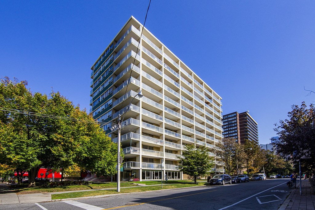 A tall building with many windows is surrounded by trees and a street with cars.