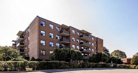 A large brick building with balconies and windows is surrounded by green bushes.