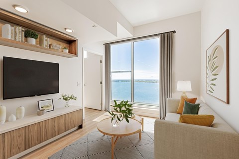 A living room with a flat screen TV, a sofa, a coffee table, and a view of the ocean through the window.