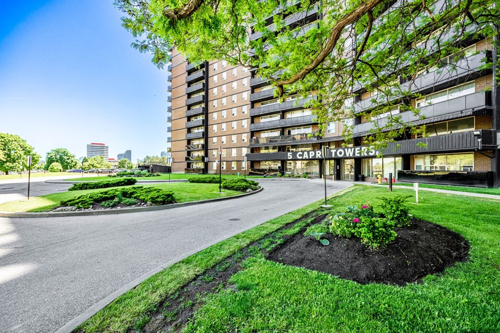 The image shows a large building with the name "CAPRI TOWERS" on it, surrounded by greenery and a clear blue sky.