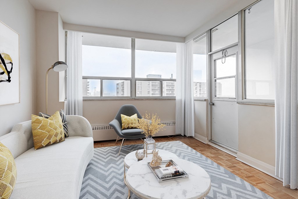 A living room with a white sofa, a grey armchair, a white coffee table, and a large window with a view of the city.