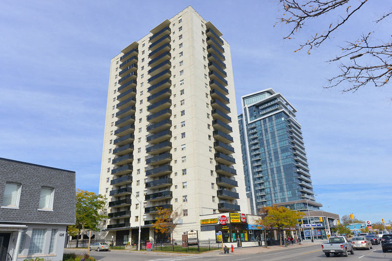 A tall beige building with balconies is in the middle of a street with cars and other buildings.