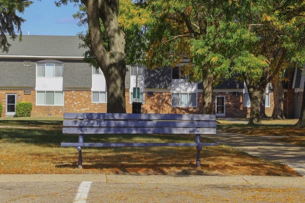 A bench sits in front of a brick building with trees in the background.