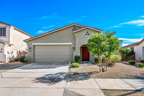 A house with a red door and a garage door is surrounded by a gravel driveway and a small tree.