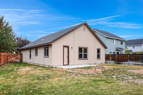 A small house with a brown door and windows is surrounded by a grassy area and a wooden fence.