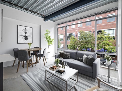 A living room with a grey couch, a grey and white coffee table, a grey dining table with grey chairs, a grey rug, a white wall with a framed picture, a window with a view of a street and buildings,