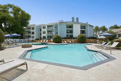 A swimming pool surrounded by lounge chairs and umbrellas in front of a multi-story apartment building.