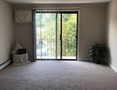 A room with a carpeted floor, a window with blinds, a white chair, a white bedspread, a black pillow, a black plant, and a black fence outside.
