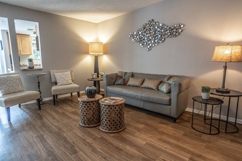 A living room with a grey couch, a chair, a table with a lamp and a plant on it, and a decorative piece on the wall.