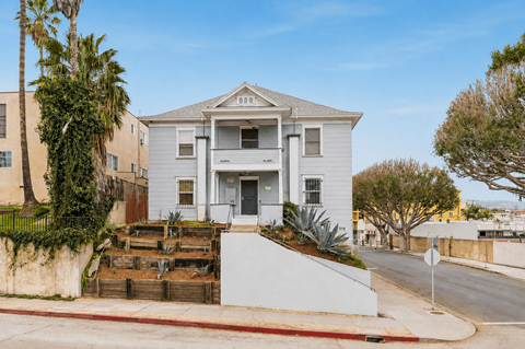 A white two-story house with a grey roof and a front yard with a wooden fence and a palm tree.