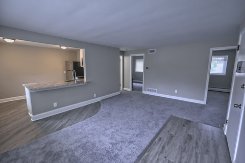 an empty living room with a kitchen and a grey carpet