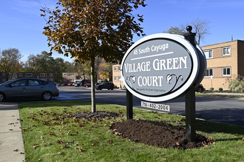 A sign for Village Green Court stands in front of a tree and a building.