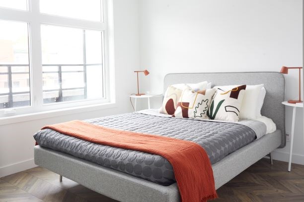 A bed with a grey blanket and orange throw is in a room with a large window.