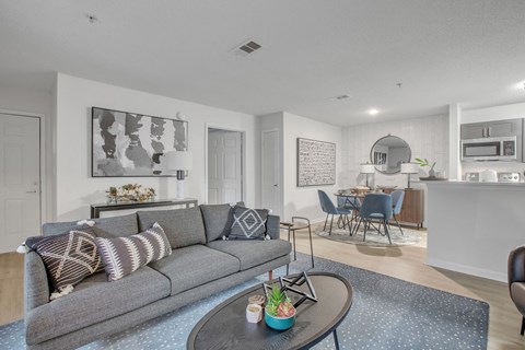 A grey couch with pillows sits in a living room with a dining area in the background.