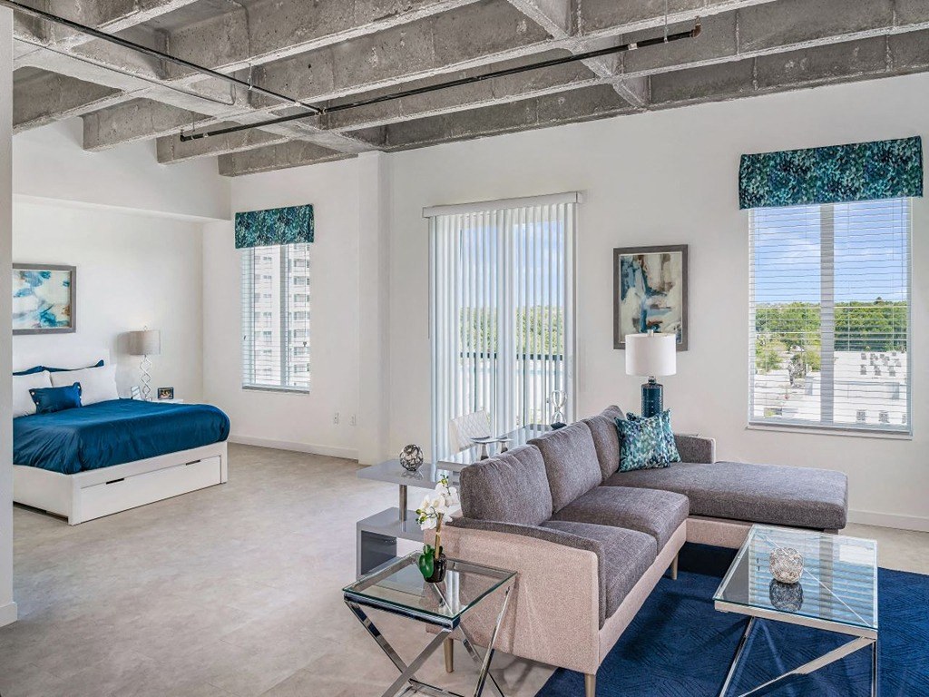A living room with a grey couch and a bed with blue bedding.