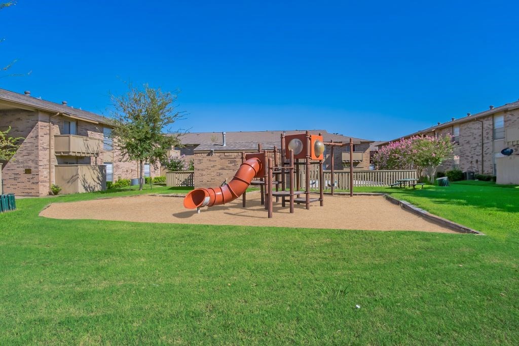 A playground with a red slide in the middle of a grassy area.