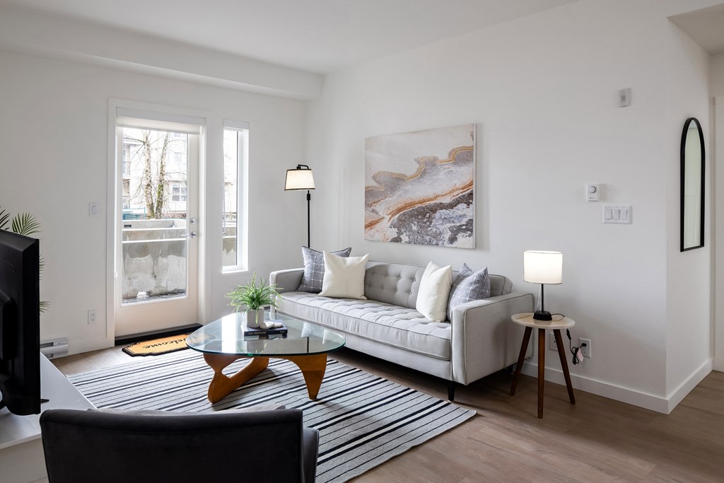 A living room with a grey couch, a black chair, a glass table, and a painting on the wall.