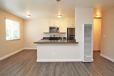 an empty living room with a kitchen with a refrigerator and a microwave