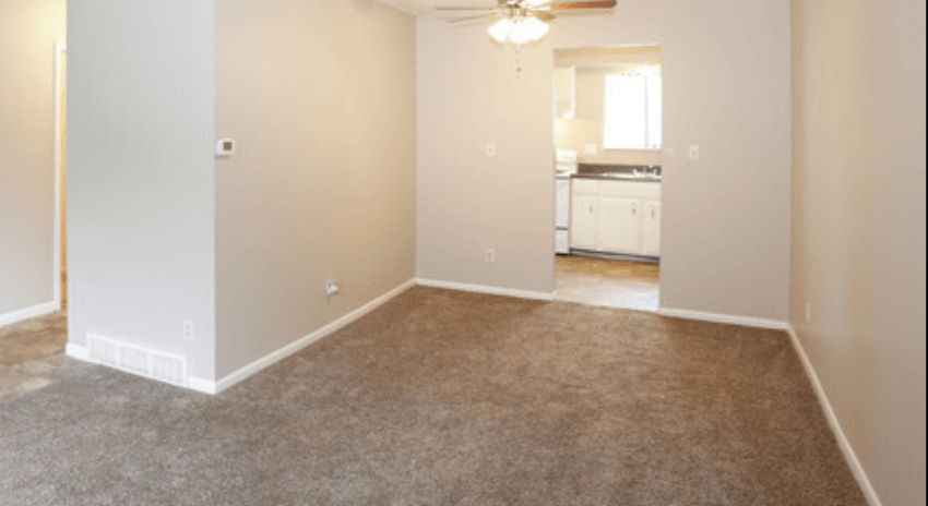 an empty living room with carpet and a kitchen in the background