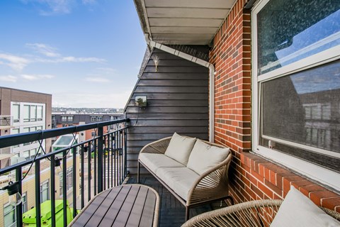 A balcony with a wicker sofa, a wooden table and a brick wall.