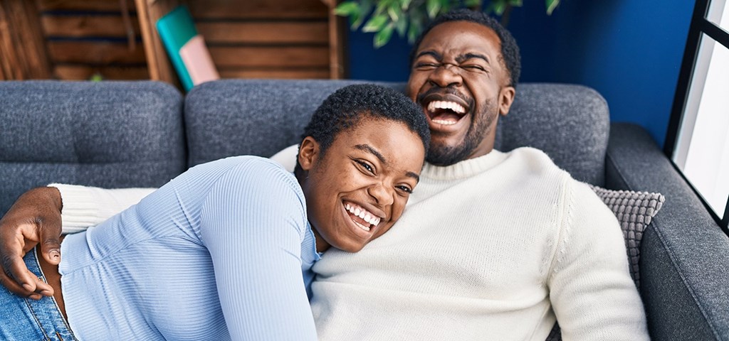 A man and a woman are laughing together on a grey couch.