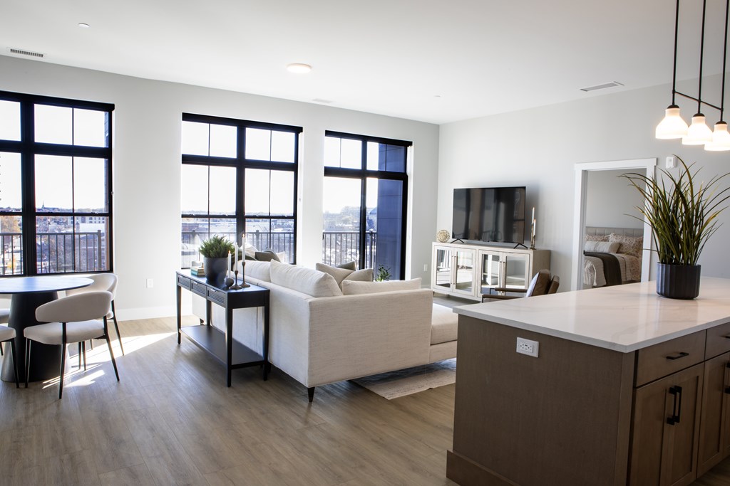 A modern living room with a white couch, a wooden cabinet, and a large window overlooking the city. at The Pearl, Fort Wayne, IN