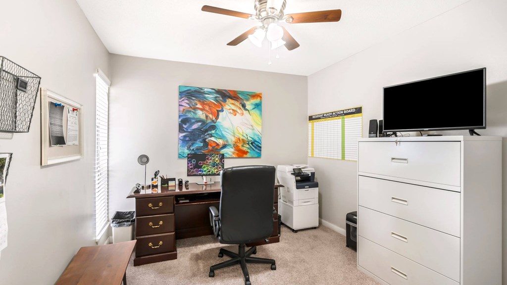 A room with a ceiling fan, a desk with a chair, and a large painting on the wall.