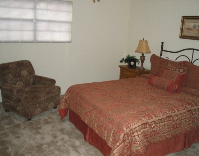A bed with a red comforter and pillows sits in a room with a chair and a lamp.